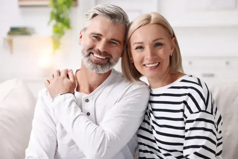 A smiling middle-aged couple with healthy teeth, sitting together in a bright, modern home.
